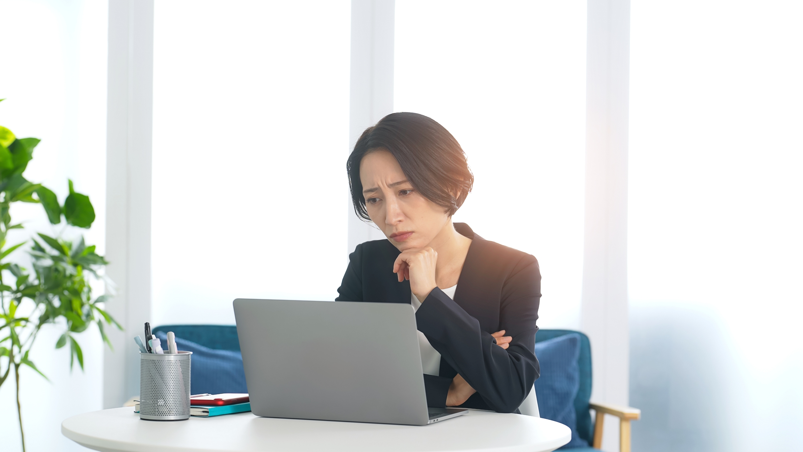 るい窓を背景に、ノートパソコンに向かうビジネスウーマンが、顎に手を添えて困惑した表情を浮かべている。仕事上の問題や課題に直面し、解決策を思案している様子