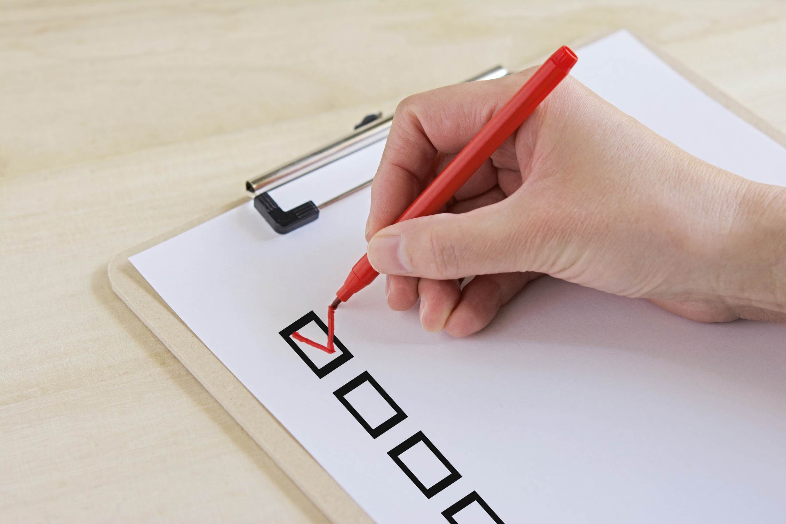 木目調の机に置かれたクリップボード上の白い書類で、赤いペンを使ってチェックボックスにチェックマークを書き込む手のクローズアップ。タスク完了や確認作業のイメージ
