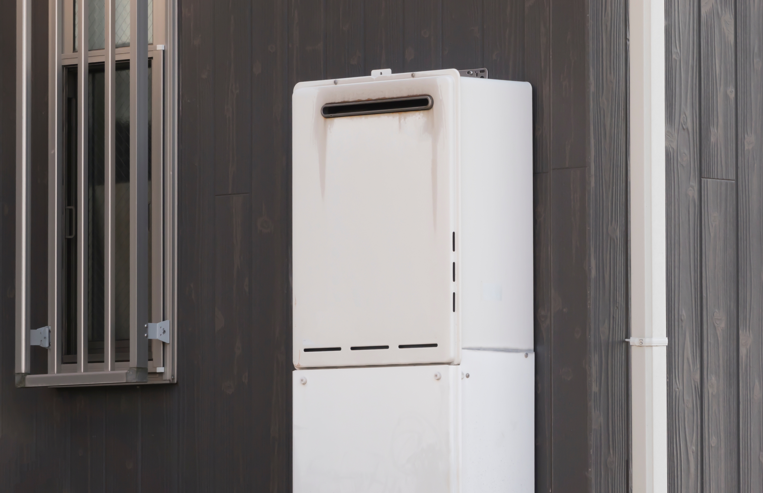 ダークグレーの木目調の外壁に設置された白い屋外給湯器。給湯器の上部排気口の下には黒い縦筋の汚れが見られる。左には防犯用の格子が付いた窓、右には白い縦樋が写っている