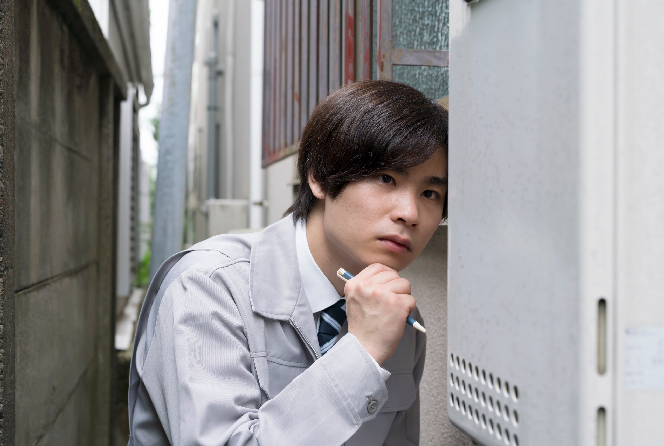 狭い路地で、グレーの作業服を着た若い男性が、屋外給湯器の隣に寄りかかり、ペンを顎に添えて真剣な眼差しで思案している。機器の点検やトラブル解決を検討している様子がうかがえる