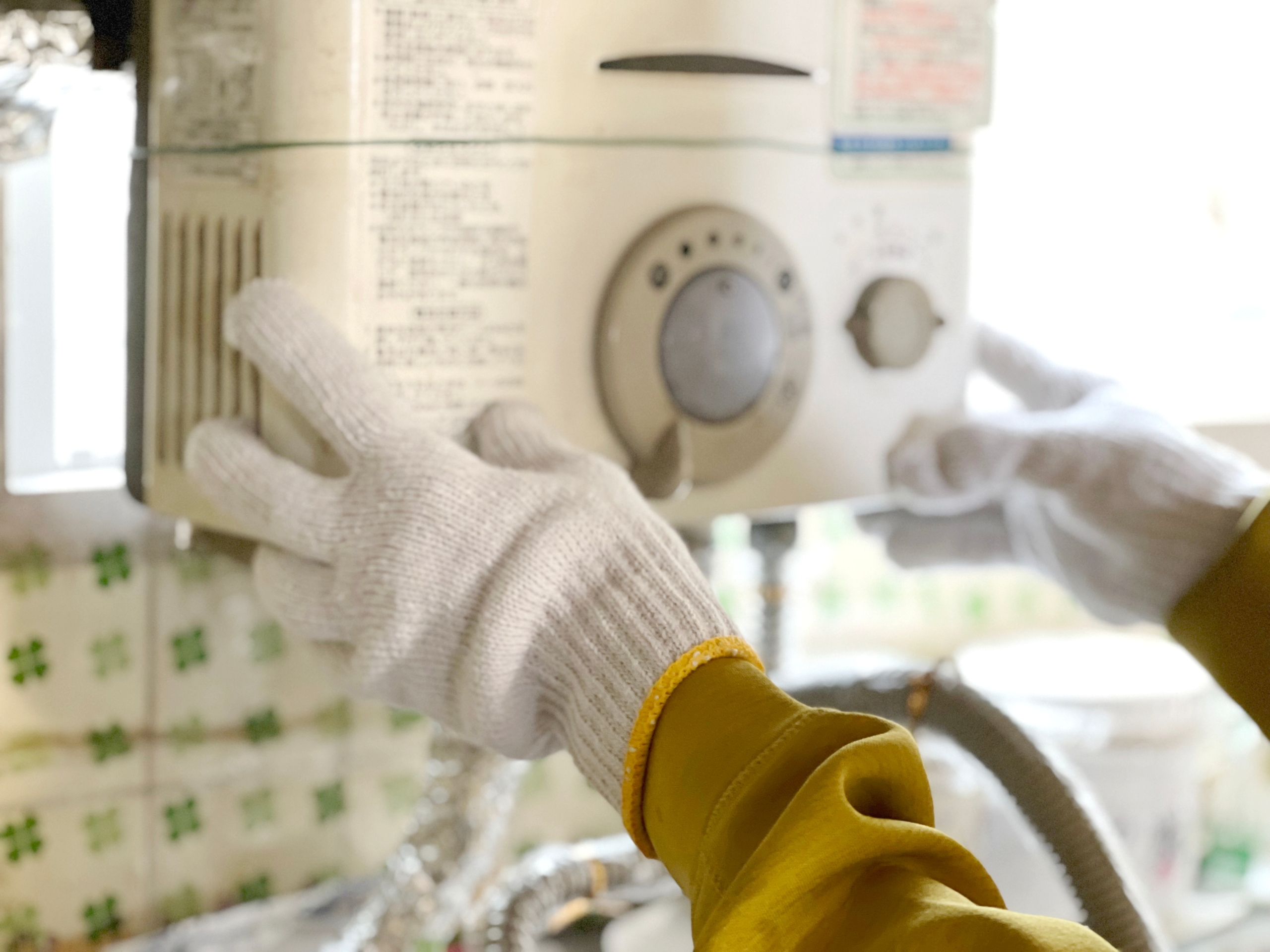 白い軍手をはめた作業員が、壁に設置された白い給湯器(または温水器)を点検している様子をクローズアップ。片手は操作パネル、もう片手は本体側面に添えられている。背景には緑色の模様入りタイルと配管の一部がぼやけて見える