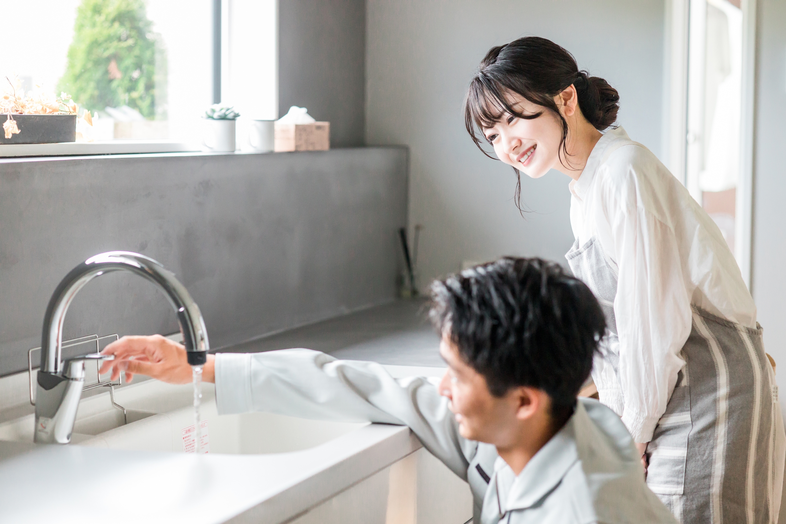 明るいキッチンで、グレーの作業服を着た男性作業員がシンクの水栓から出る水を点検している。隣にはエプロン姿の女性が笑顔でその様子を見守っている。水回り設備の点検や修理の様子を表す