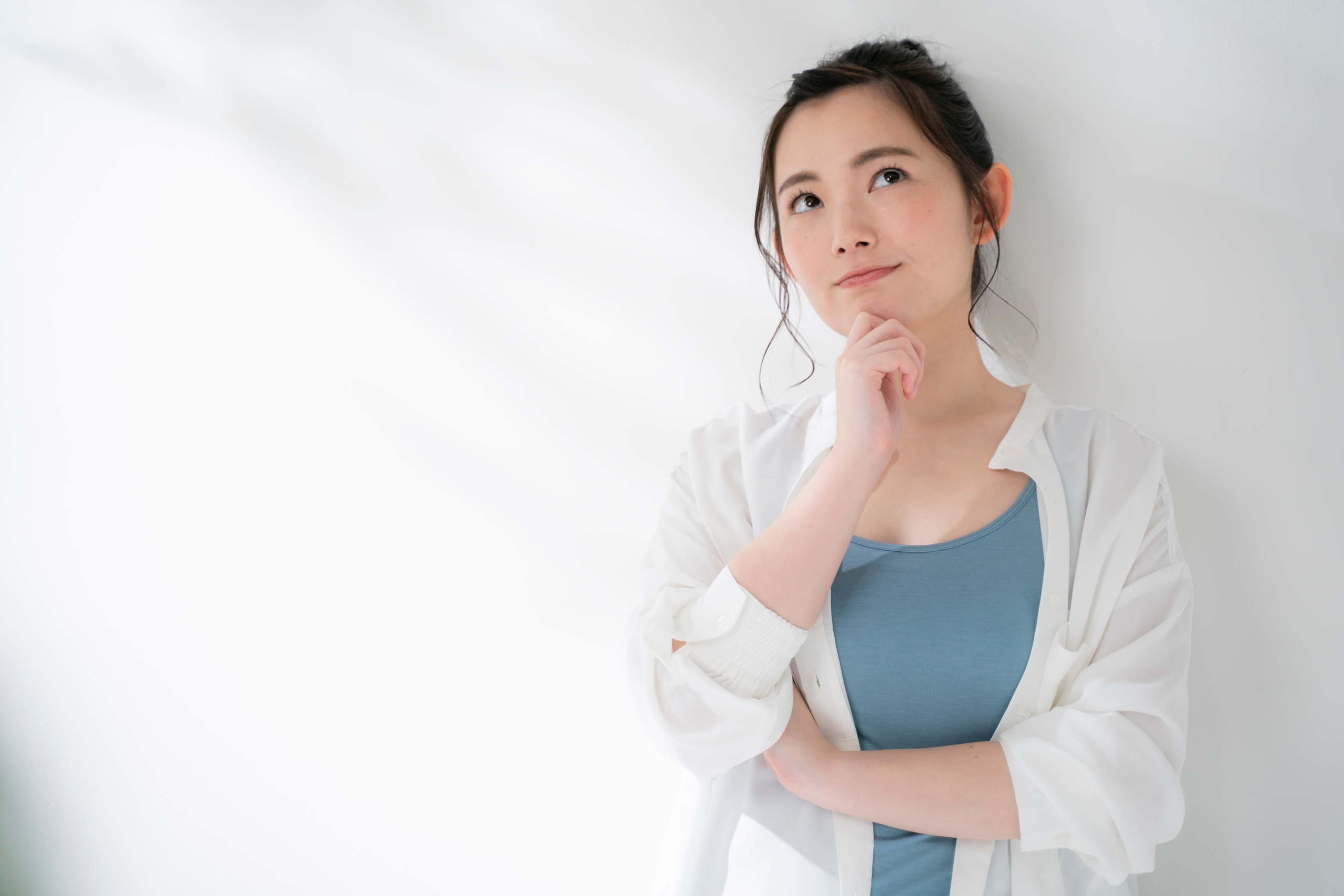 白い壁の前で顎に手を当て、何かを考えて上を見上げる若い女性。ブルーのインナーに白いシャツを羽織り、左側から木漏れ日のような影が差し込んでいます